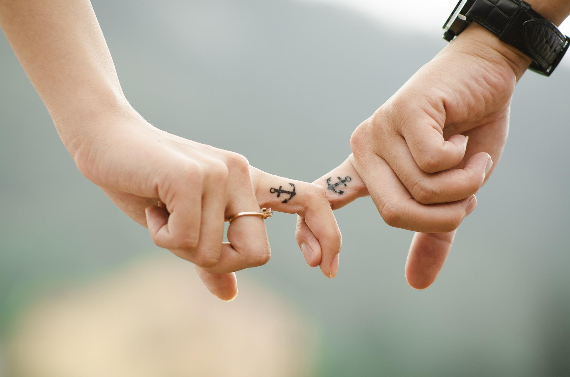 Une mais féminine et une main masculine se tiennent par le doigt, chacune des deux a un tatouage représentant un ancre de marine